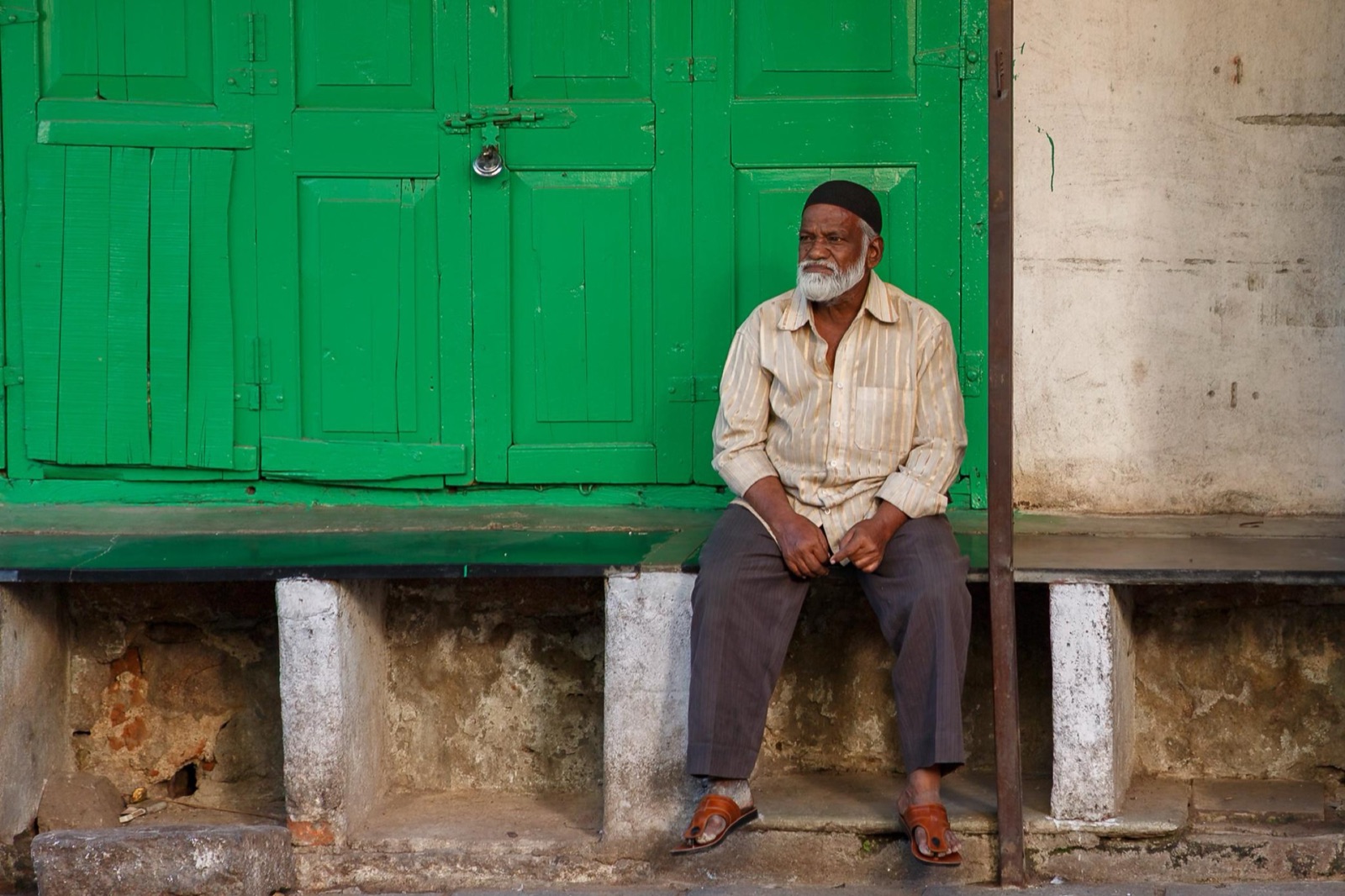 An older man in a cap sitting on a stone ledge in front of bright green wooden doors.