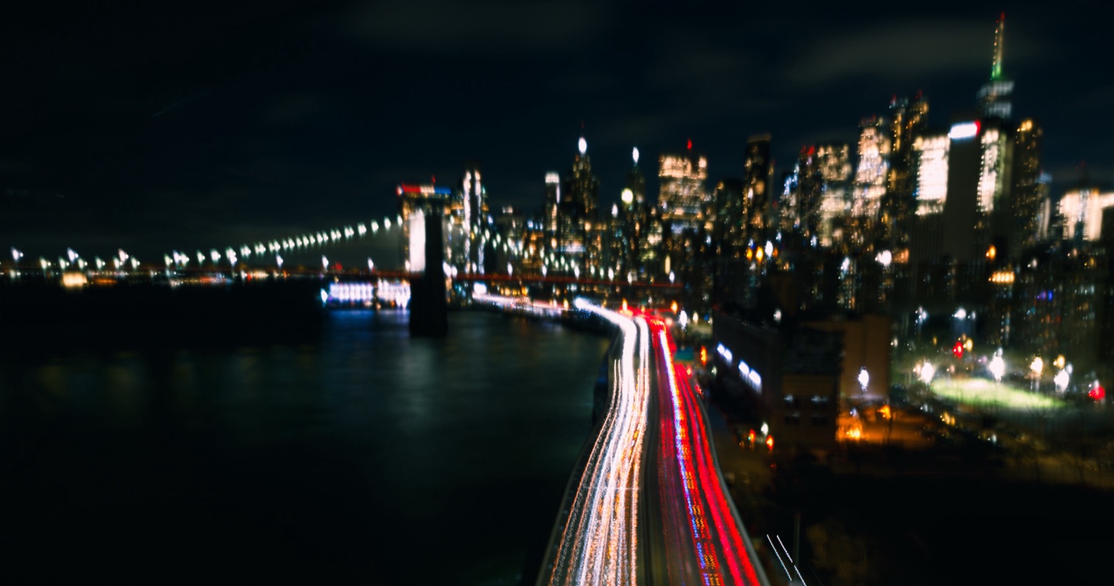 A city skyline at night across a river, with red and white light trails from traffic along the highway.