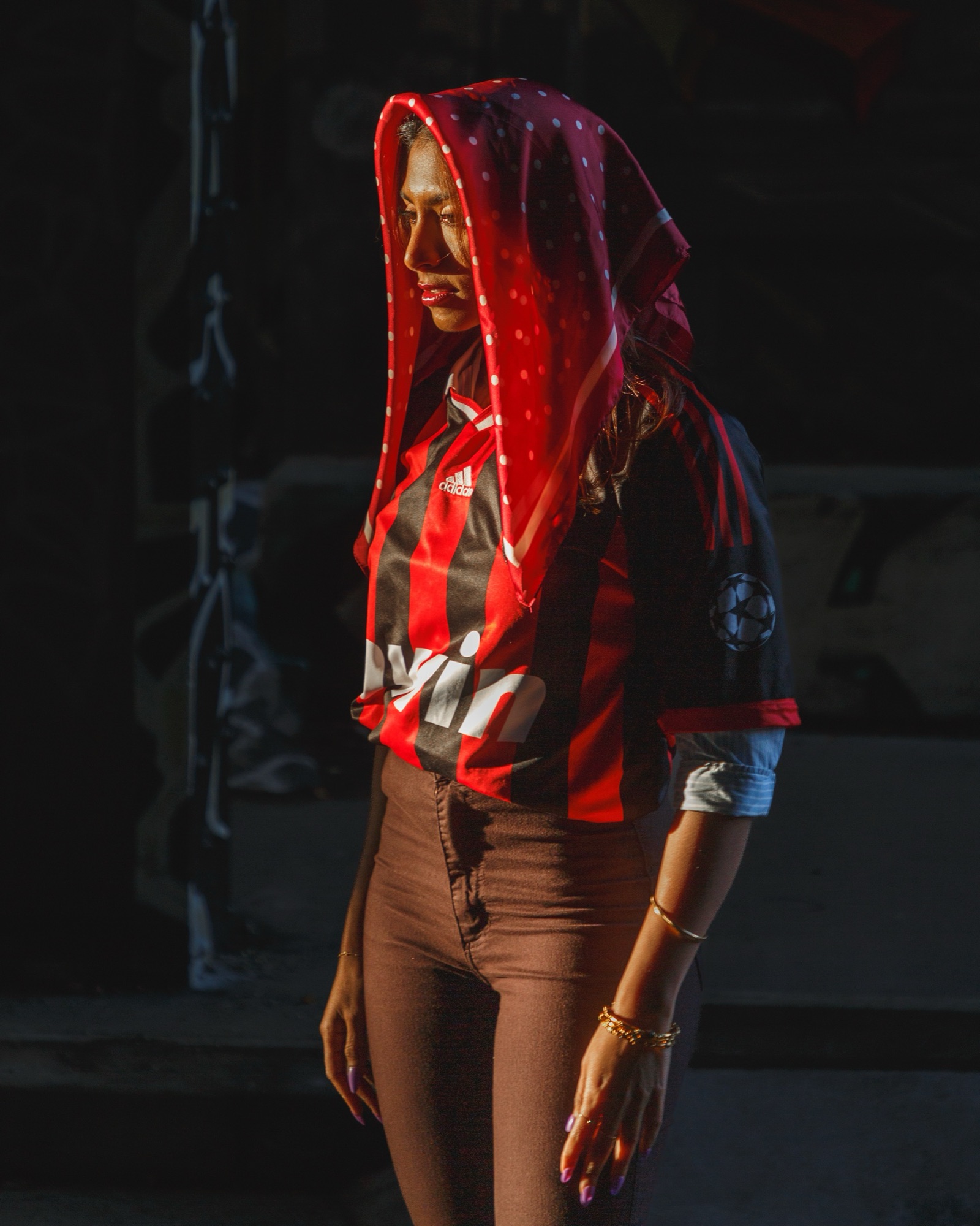 A woman in a red headscarf and red-and-black football jersey, lit by a shaft of sunlight against a dark wall.