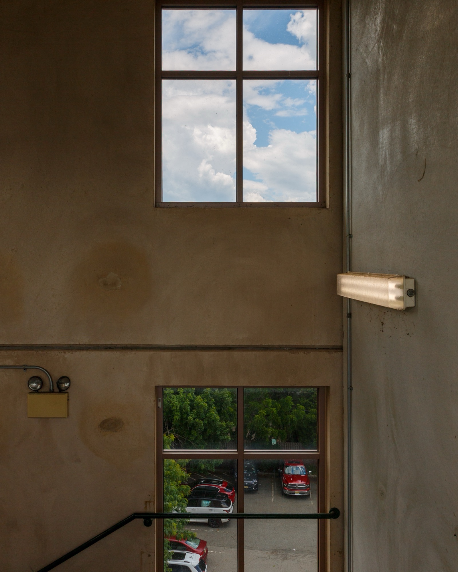 A dim stairwell with two windows, blue sky in the upper one and parked cars below.