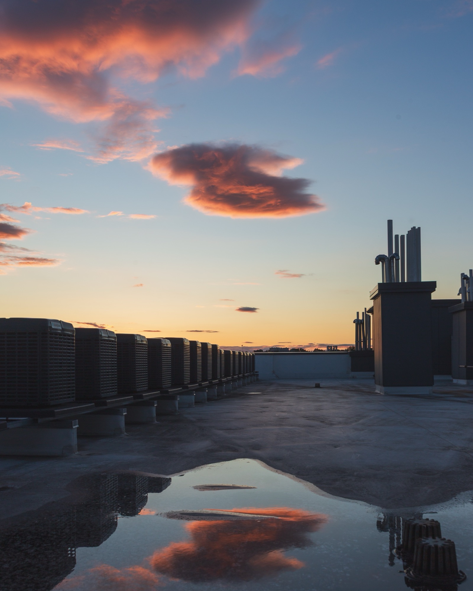 A rooftop at dusk, pink clouds reflected in a puddle between rows of air-conditioning units.