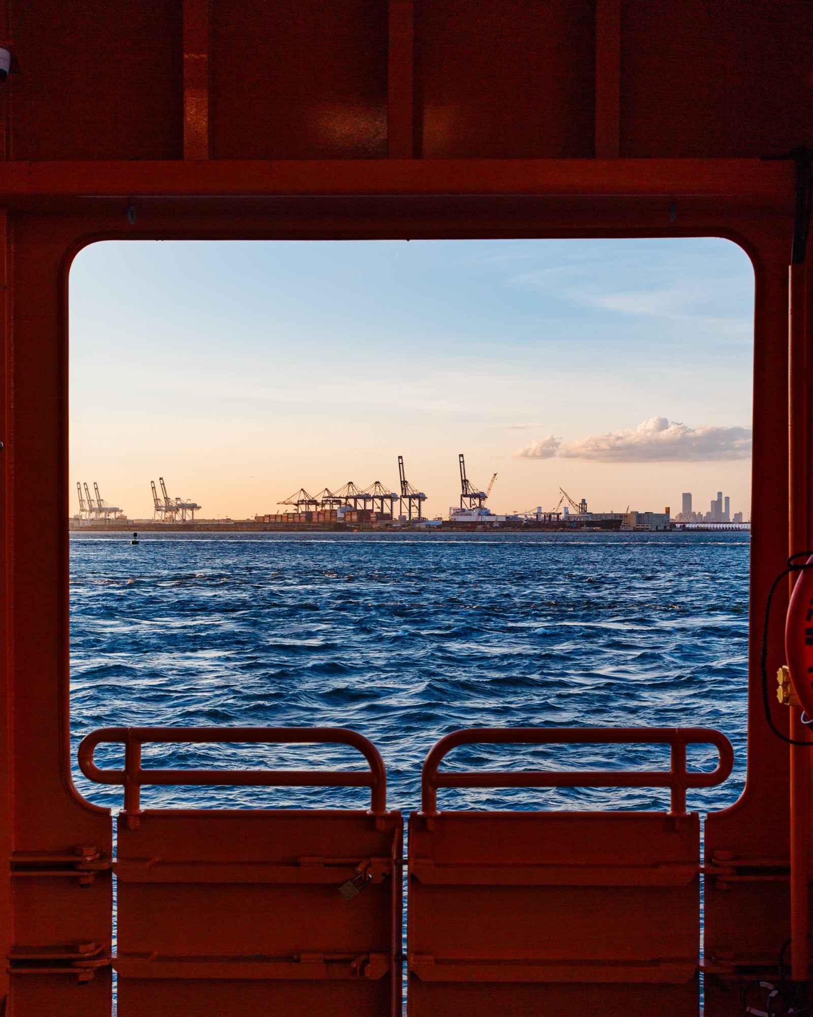 View through the red-framed window of a ferry, looking across blue water to cranes on the far shore.