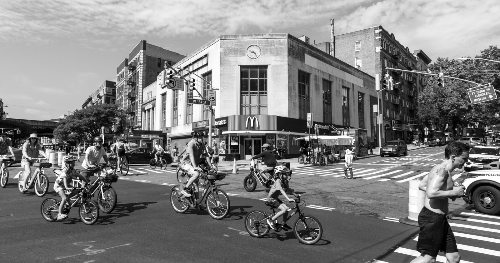 Black-and-white street scene of children riding bikes through a busy city intersection.