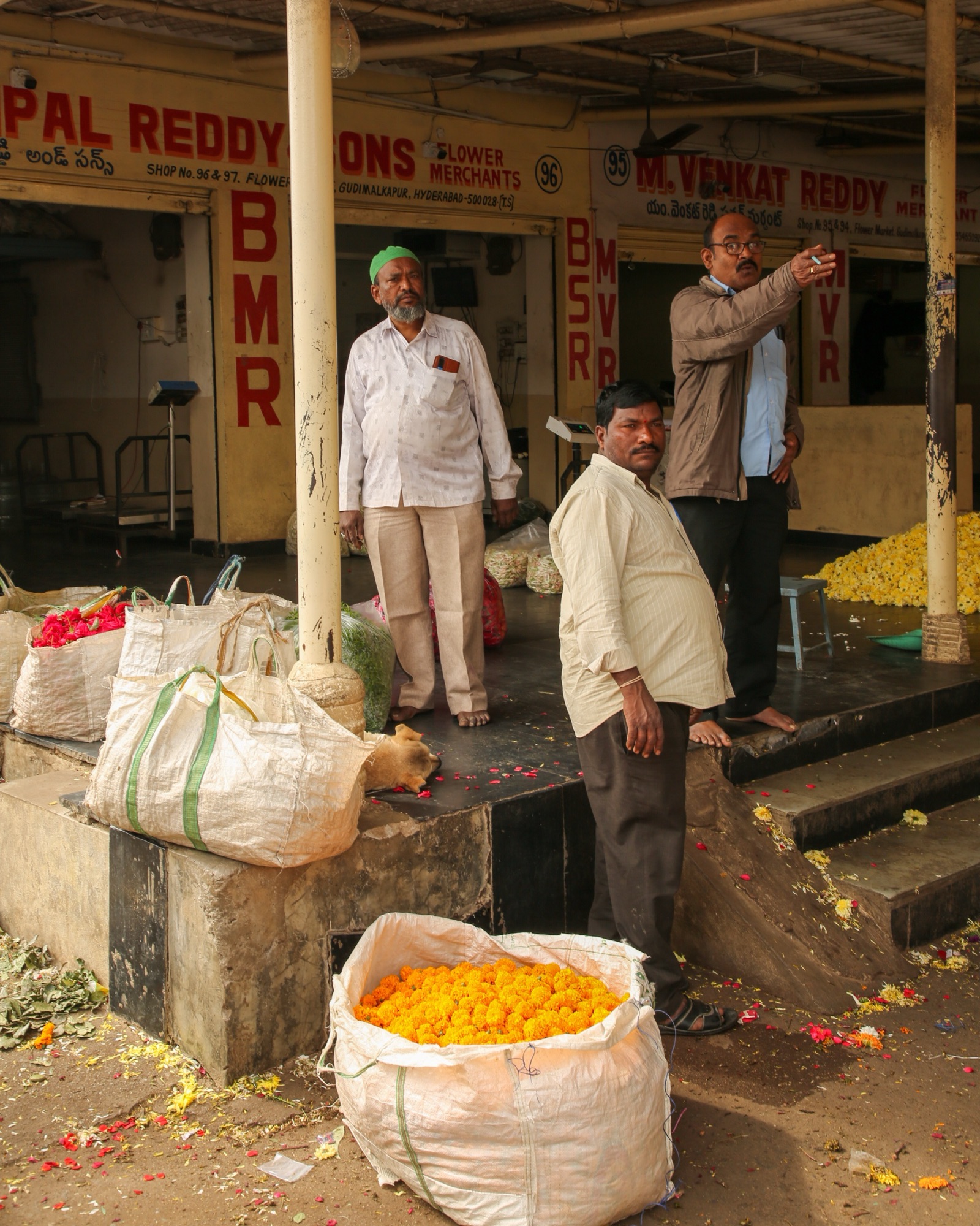 Three men standing among sacks of marigolds at a flower market stall.
